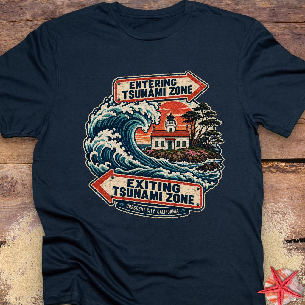 A navy blue short-sleeve t-shirt featuring a vintage-style graphic of a lighthouse and tsunami wave with directional arrows reading 'ENTERING TSUNAMI ZONE' and 'EXITING TSUNAMI ZONE' with 'Crescent City, California' below.