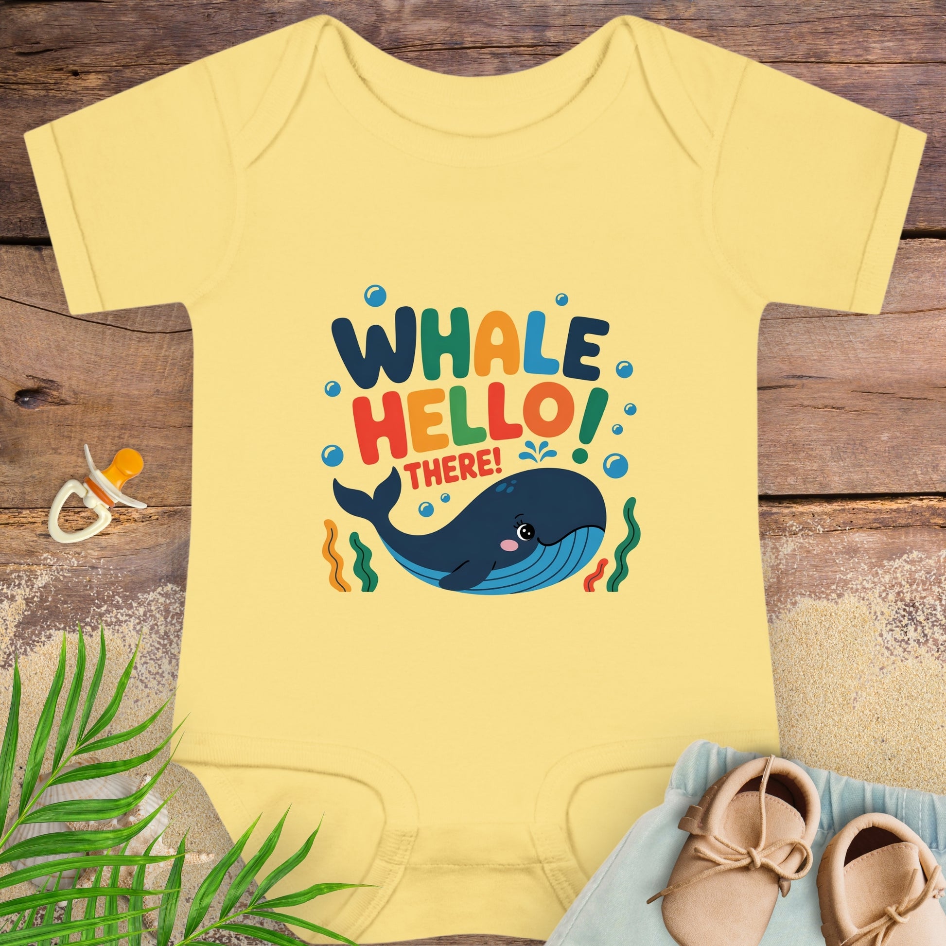 Yellow baby onesie with whale design and text on a wooden surface with shoes and leaves.