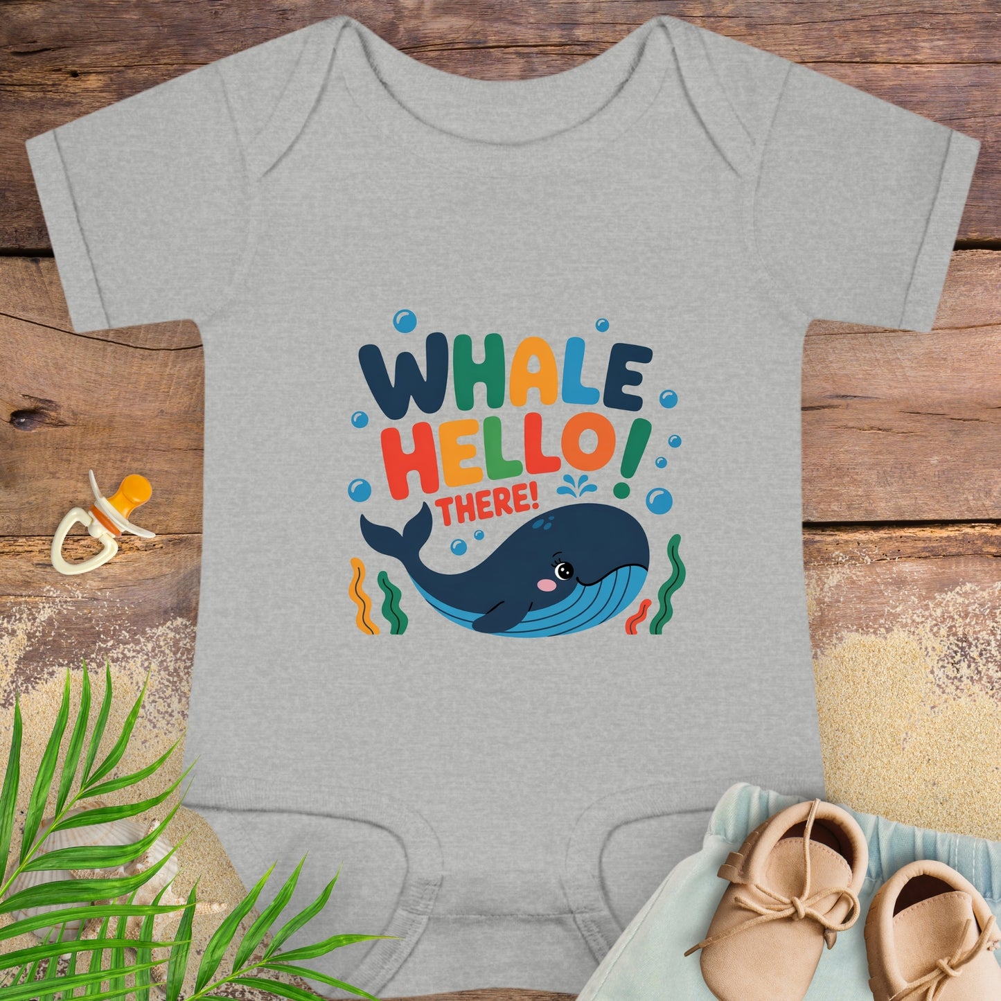 Gray baby onesie with colorful 'Whale Hello! There!' design on a wooden surface with shoes and a pacifier.