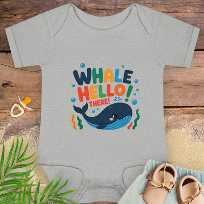 Gray baby onesie with colorful 'Whale Hello! There!' design on a wooden surface with shoes and a pacifier.