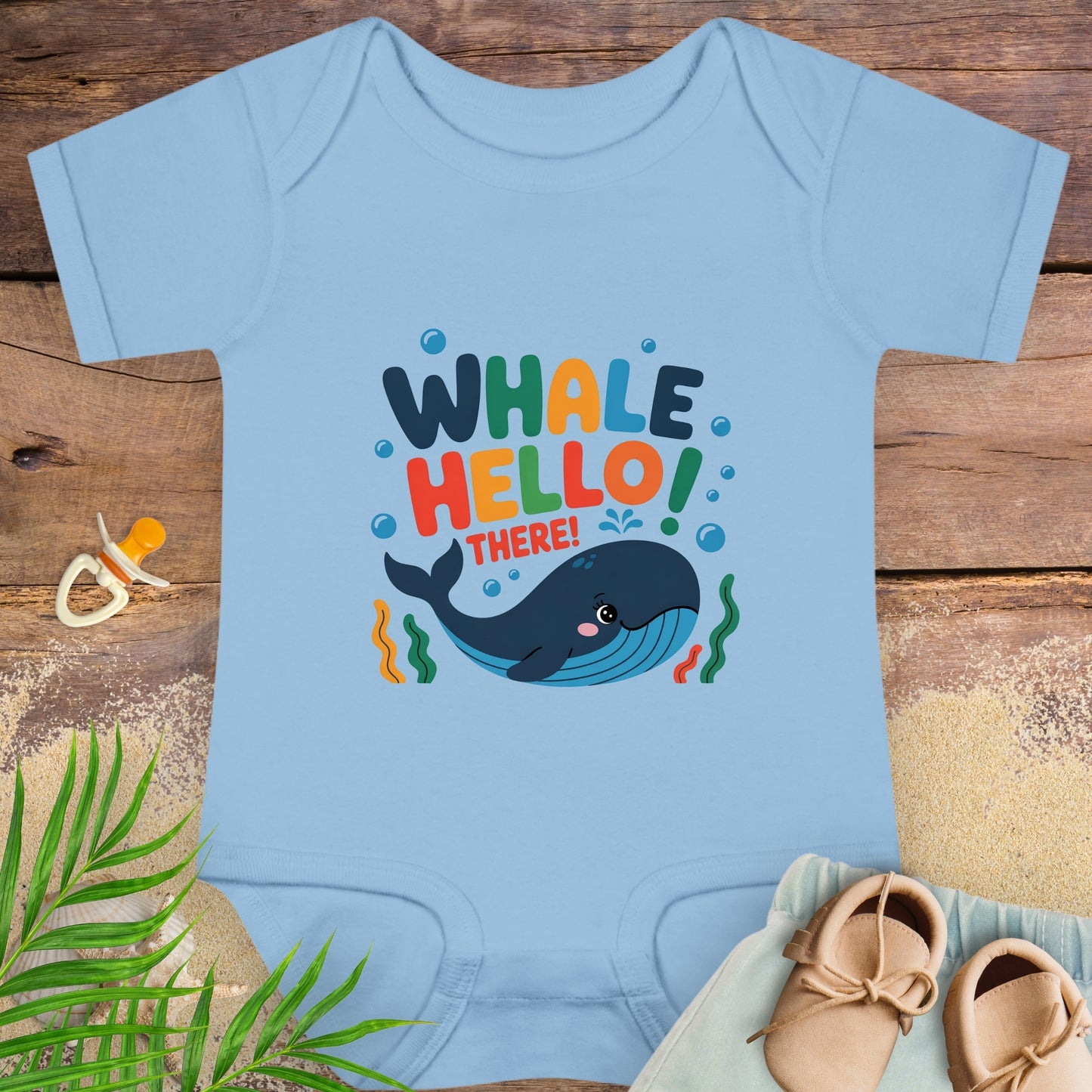 Light blue baby onesie with whale graphic and text on a wooden surface with shoes and a pacifier.
