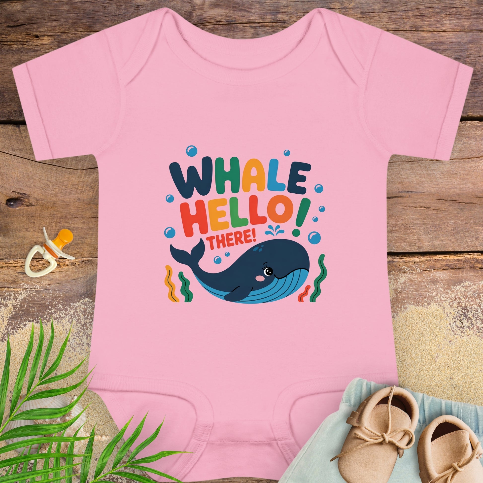 Pink baby onesie with whale design and text on a wooden surface with shoes and leaves.