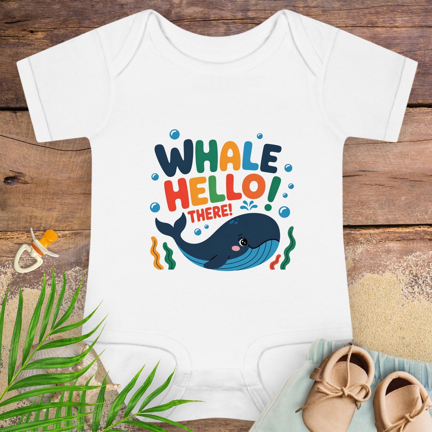 White baby onesie with colorful 'Whale Hello! There!' design on a wooden surface with baby items.